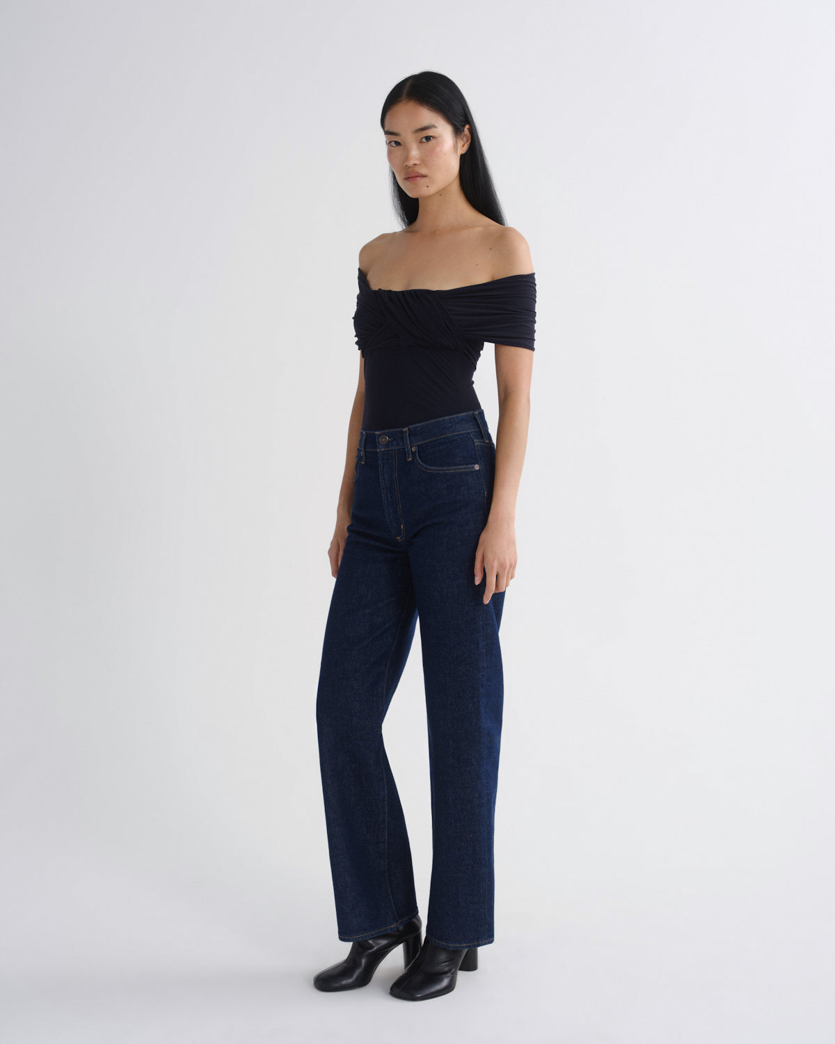 A model wearing a black off-the-shoulder ruched top, dark blue wide-leg jeans, and black leather boots, standing against a plain light gray studio background.