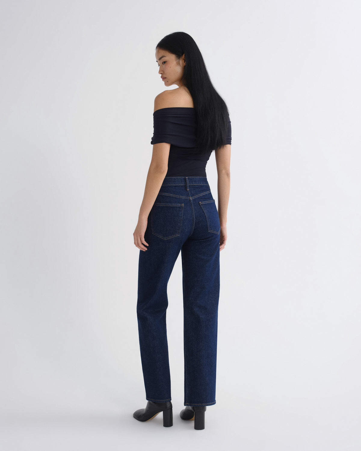 A woman with long black hair is shown from the back wearing dark blue high-waisted straight-leg jeans, a dark navy off-the-shoulder top, and black heeled boots against a plain light gray studio background.