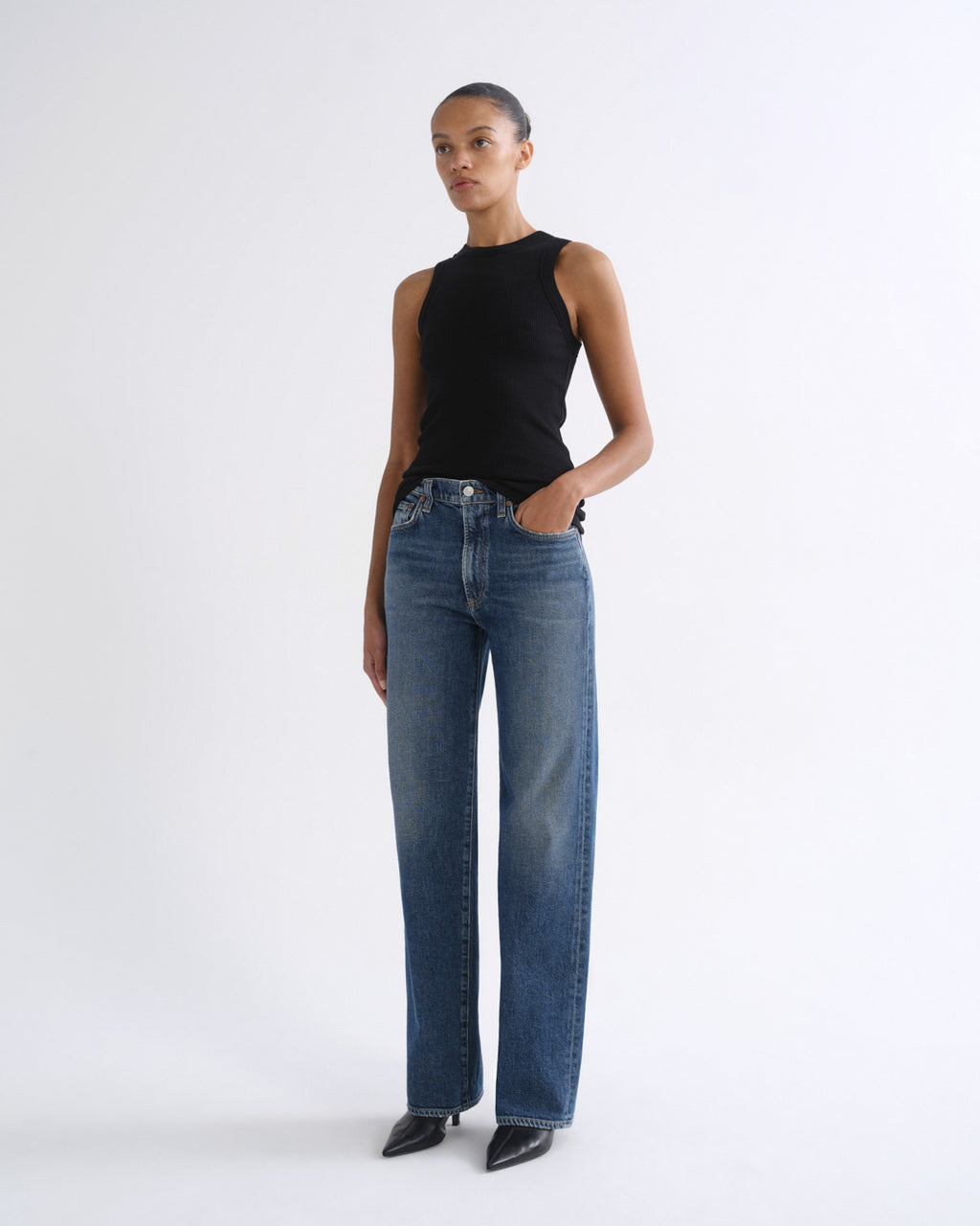A model in profile wearing a black sleeveless top and wide-leg blue jeans, standing against a plain light gray studio background.