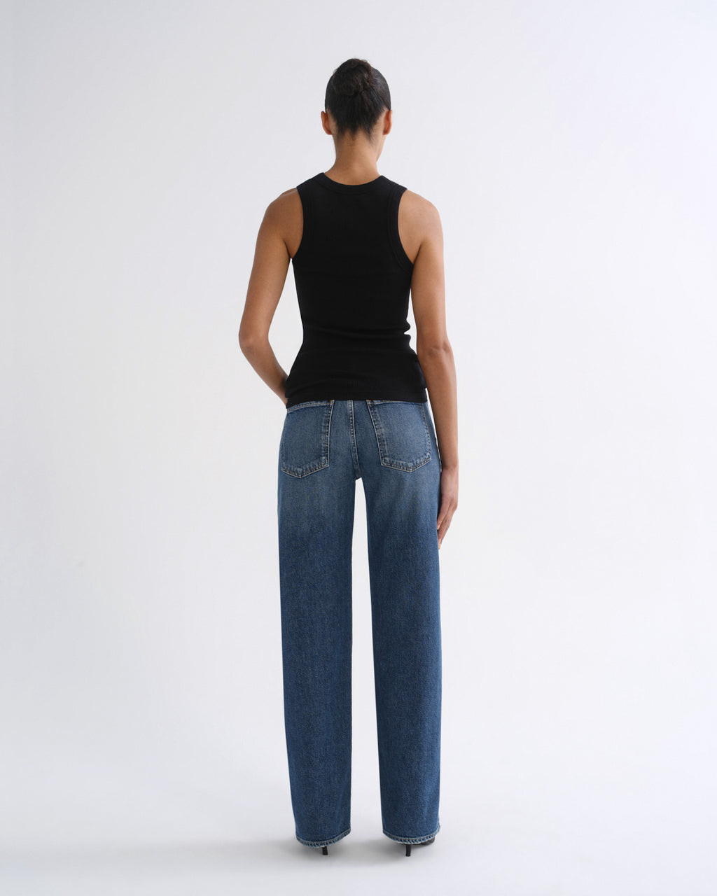 A model viewed from the back wearing a black sleeveless top and wide-leg blue jeans against a plain light gray studio background.
