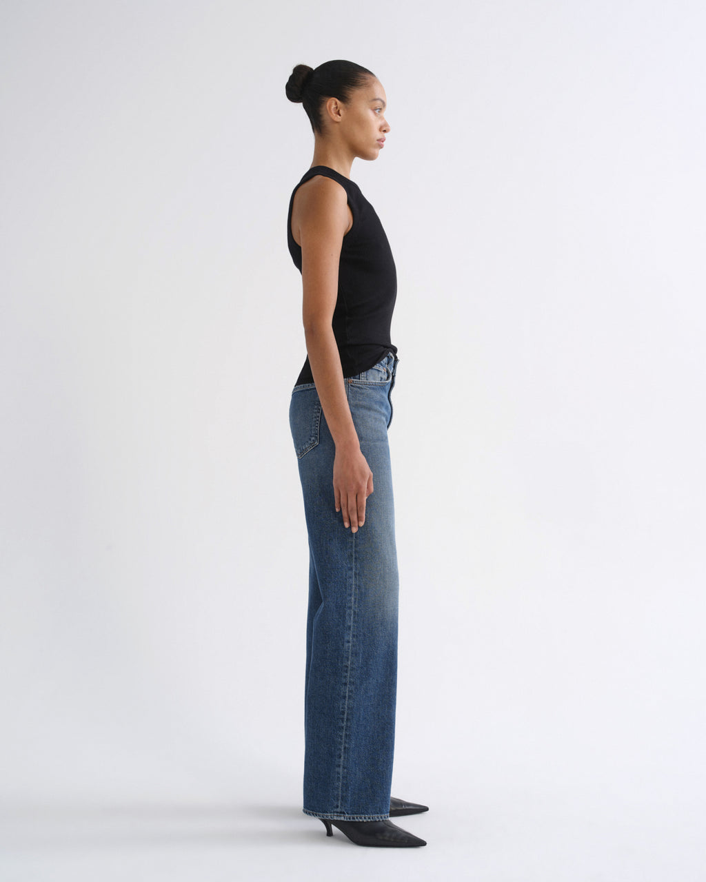 A model in profile wearing a black sleeveless top and wide-leg blue jeans, standing against a plain light gray studio background.