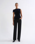 A model stands against a plain white studio background wearing a dark navy velvet off-the-shoulder top and black wide-leg trousers.