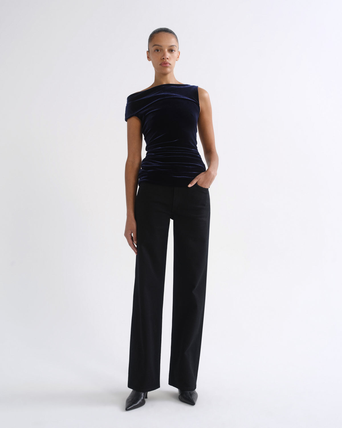 A model stands against a plain white studio background wearing a dark navy velvet off-the-shoulder top and black wide-leg trousers.