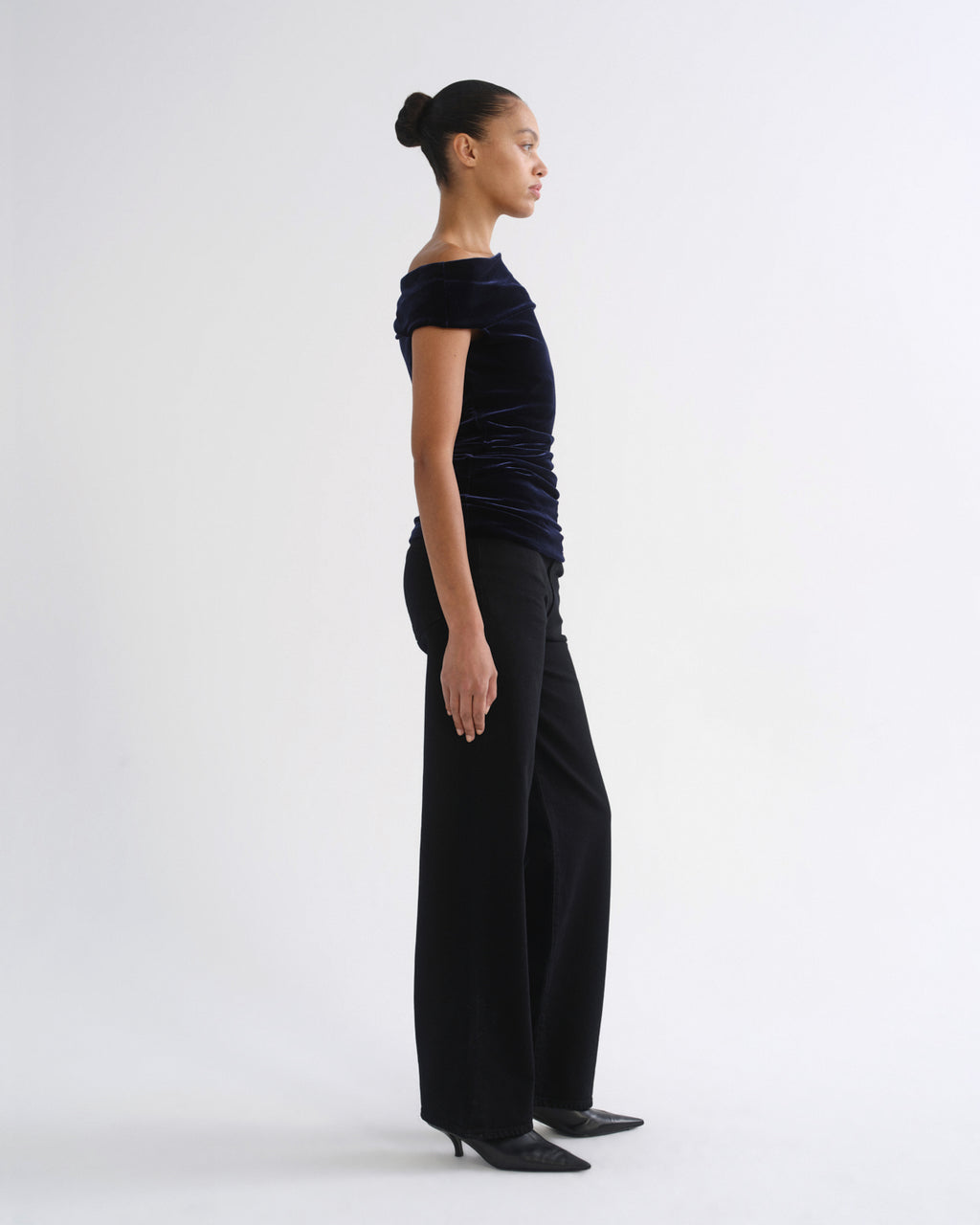A model in profile wearing a dark navy off-the-shoulder velvet top and black wide-leg trousers, standing against a plain light gray studio background.