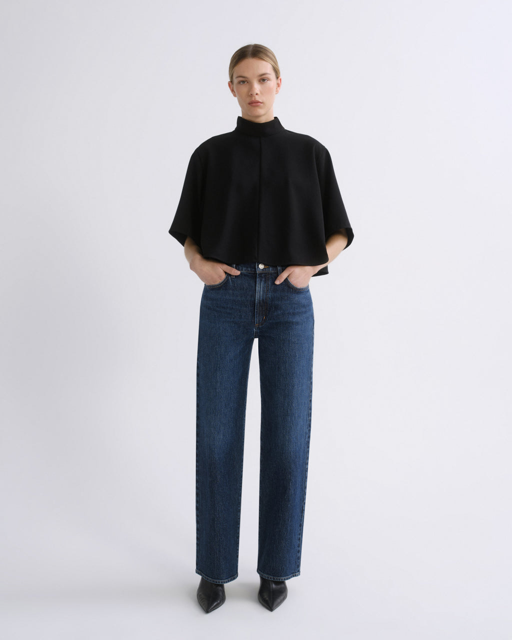 Woman wearing a black cape sleeve top with medium blue wide leg jeans on a grey background.