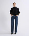 Woman wearing a black cape sleeve top with medium blue wide leg jeans on a grey background.