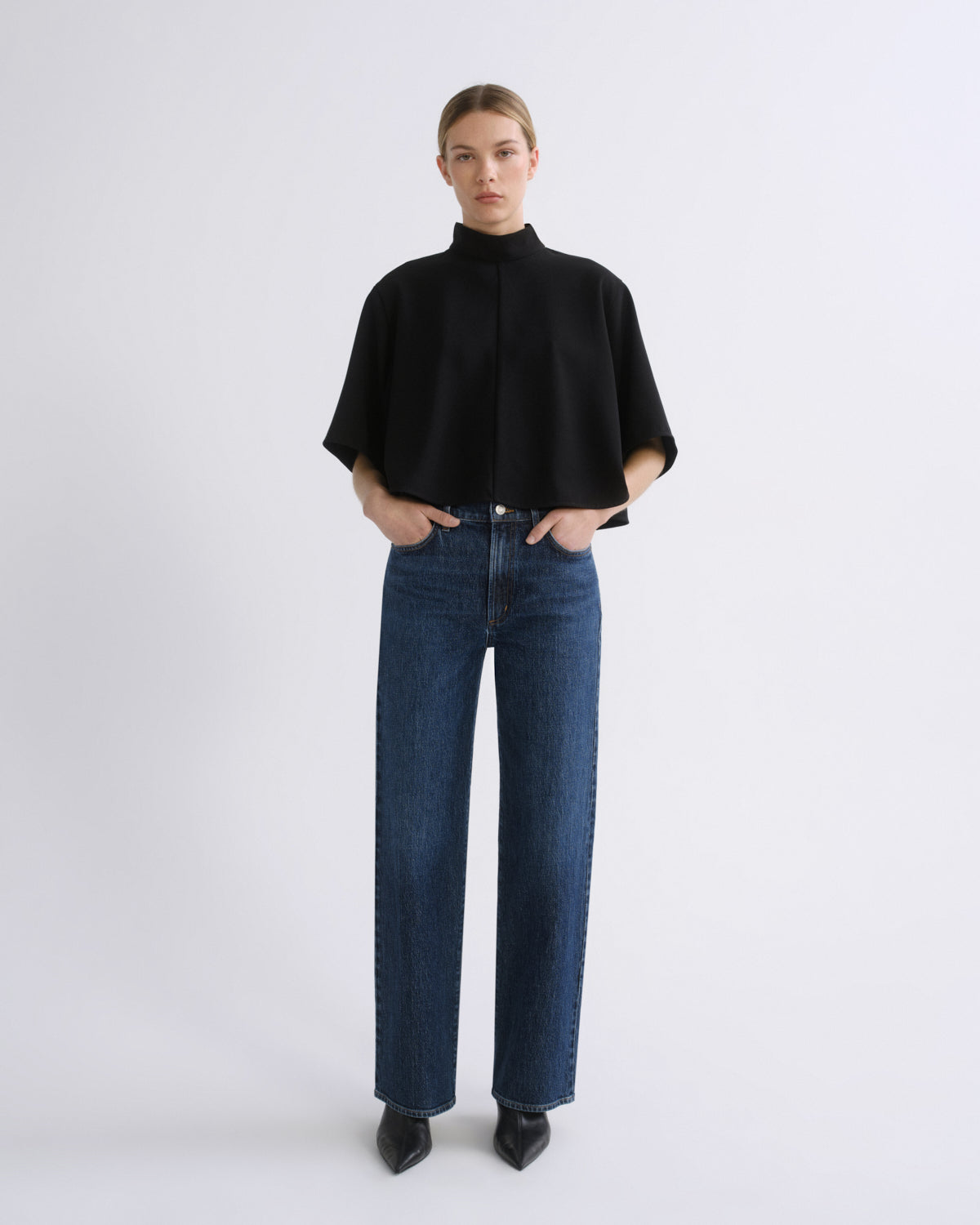 Woman wearing a black cape sleeve top with medium blue wide leg jeans on a grey background.