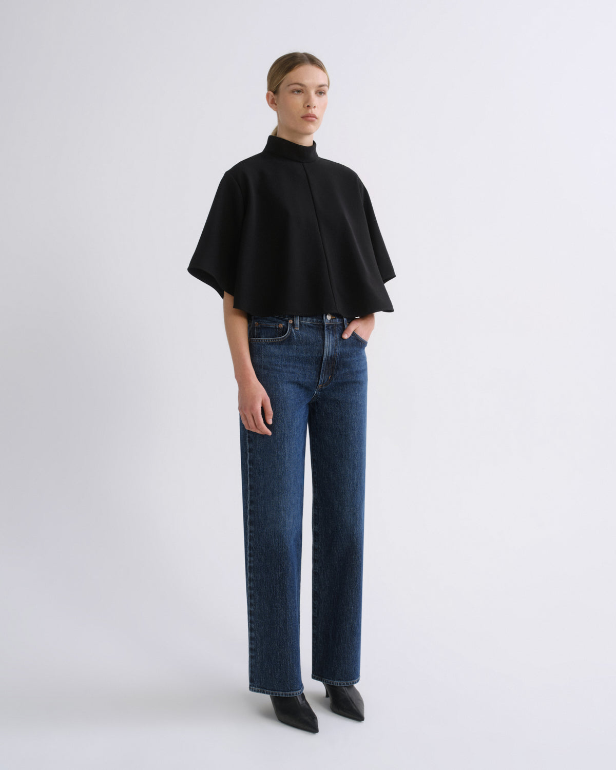 Woman wearing a black cape sleeve top with medium blue wide leg jeans on a grey background.