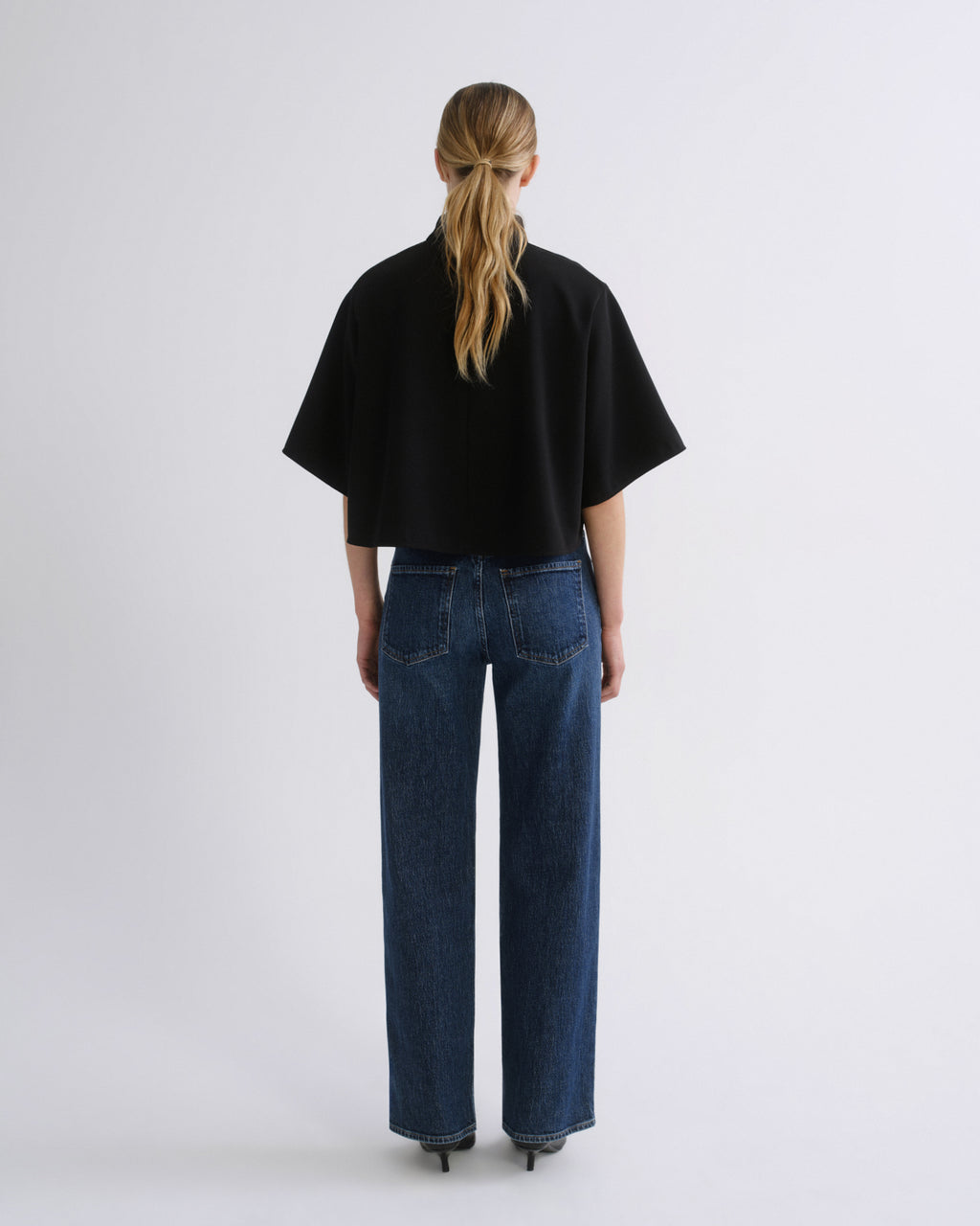 Woman wearing a black cape sleeve top with medium blue wide leg jeans viewed from the back on a grey background.