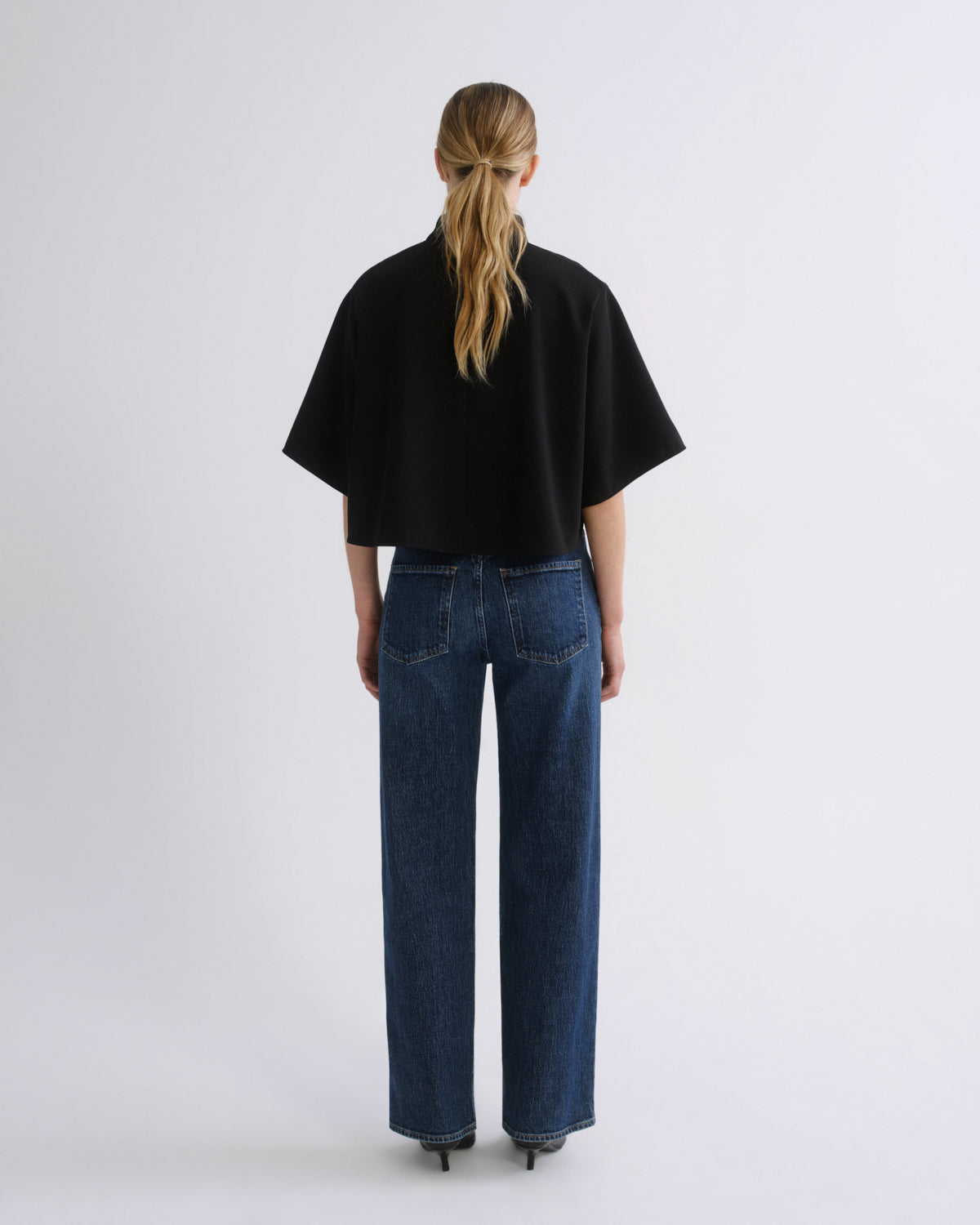 Woman wearing a black cape sleeve top with medium blue wide leg jeans viewed from the back on a grey background.