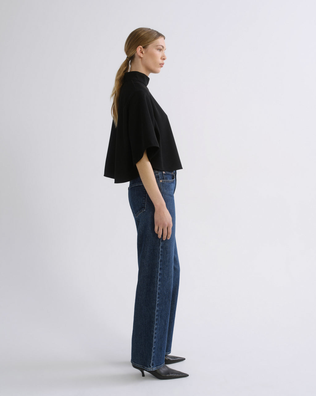 Woman wearing a black cape sleeve top with medium blue wide leg jeans viewed from the side on a grey background.