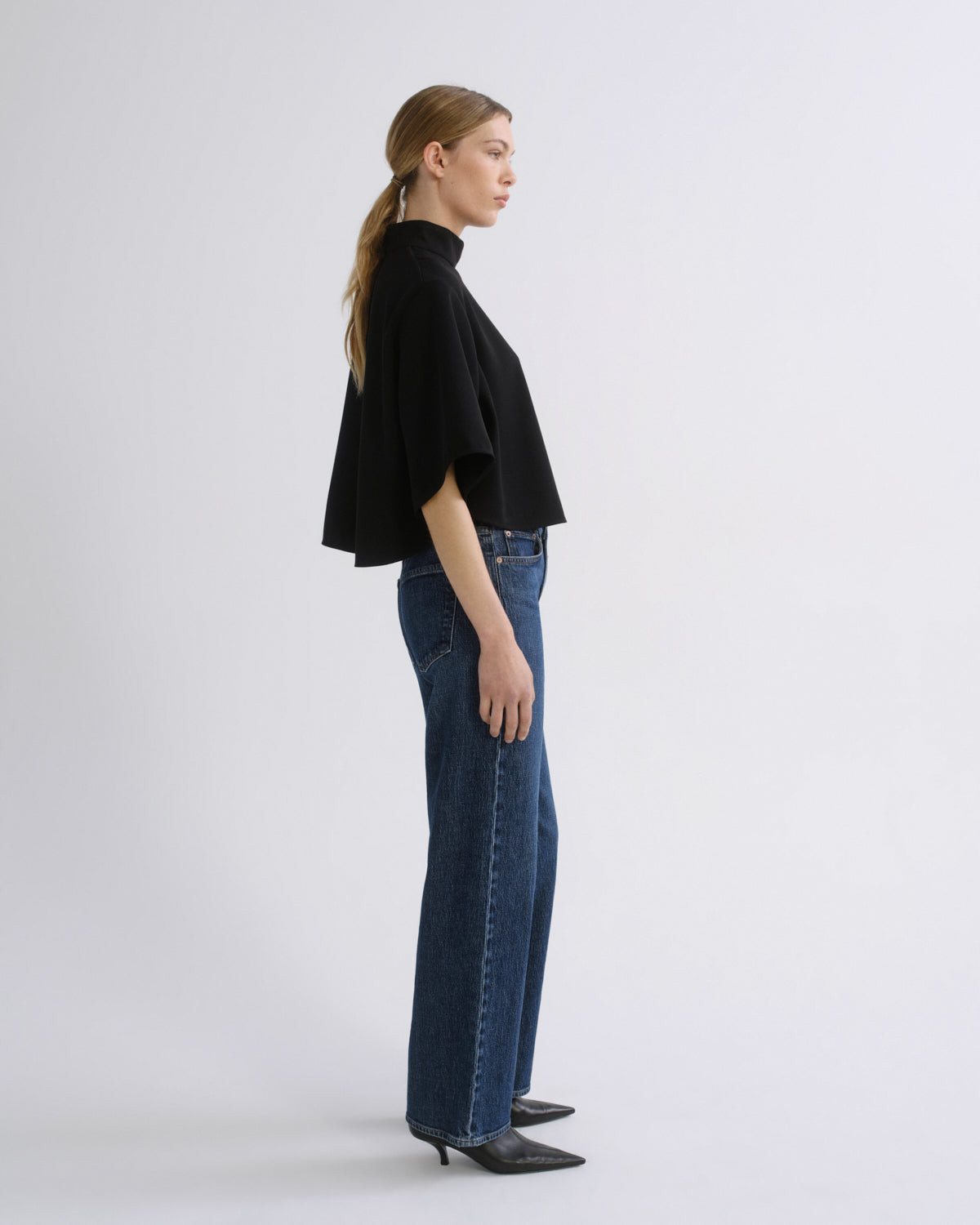 Woman wearing a black cape sleeve top with medium blue wide leg jeans viewed from the side on a grey background.