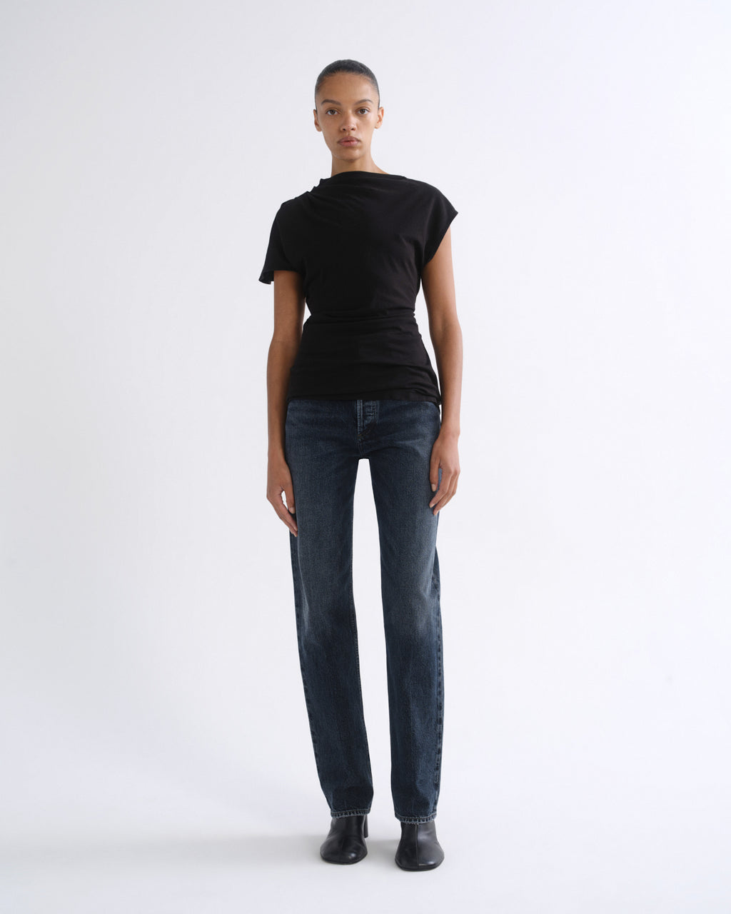 Woman wearing a black short sleeve top with dark indigo straight leg jeans on a grey background.