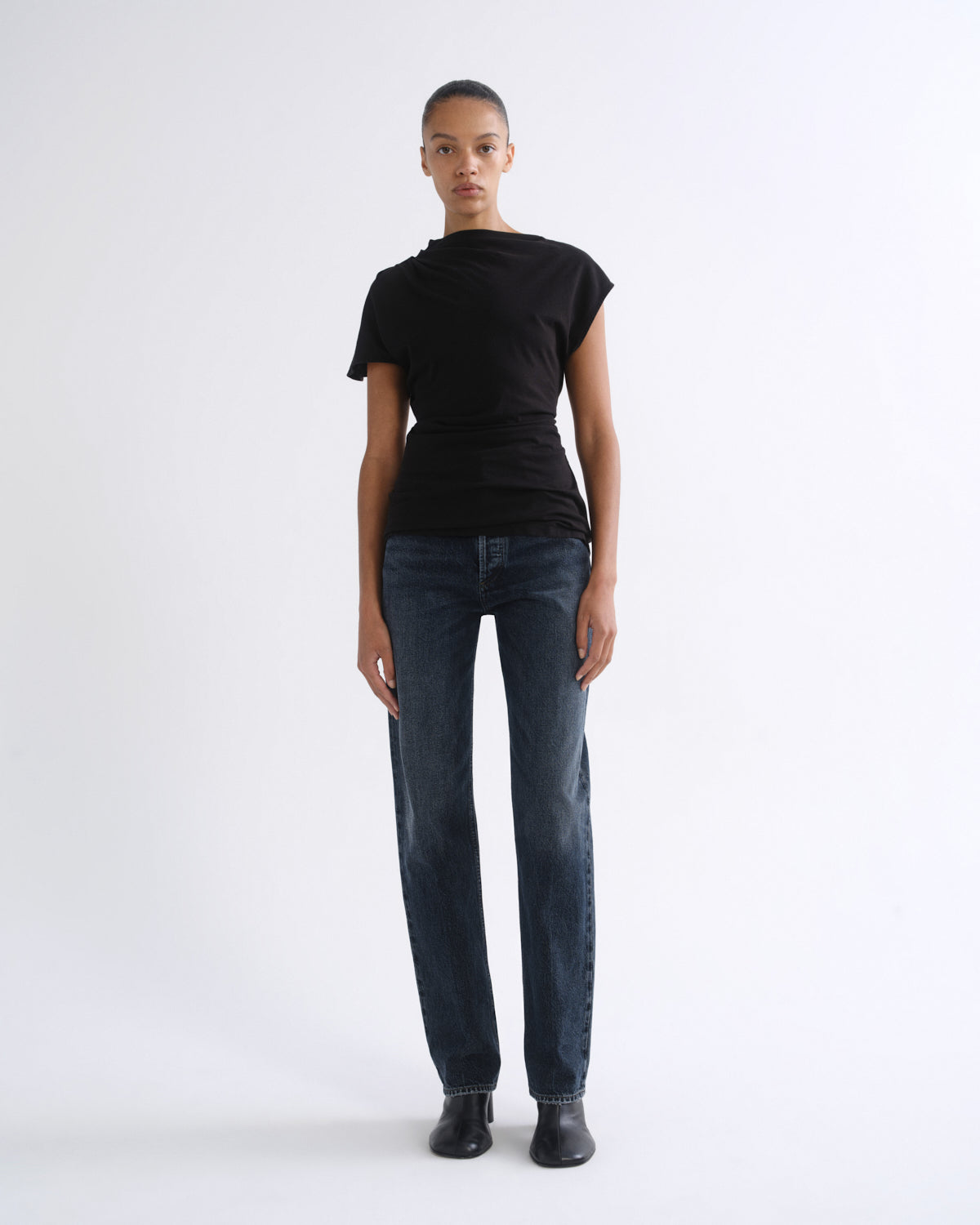 Woman wearing a black short sleeve top with dark indigo straight leg jeans on a grey background.