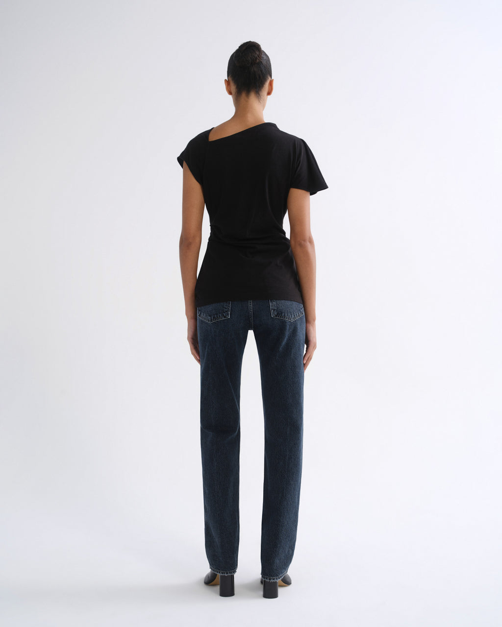 Woman wearing a black short sleeve top with dark indigo straight leg jeans viewed from the back on a grey background.