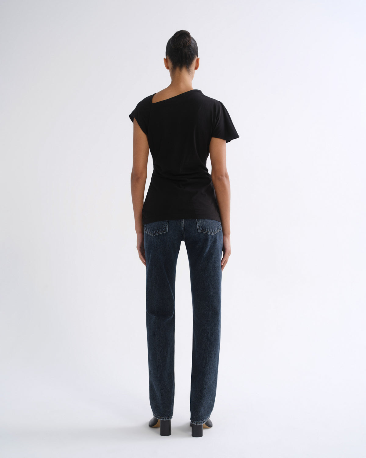 Woman wearing a black short sleeve top with dark indigo straight leg jeans viewed from the back on a grey background.