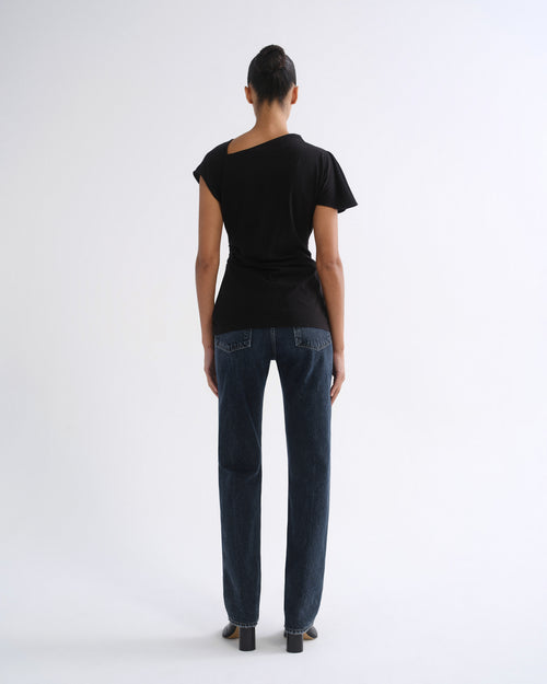 Woman wearing a black short sleeve top with dark indigo straight leg jeans viewed from the back on a grey background.