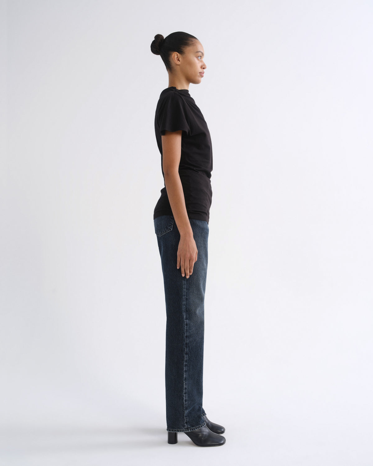 Woman wearing a black short sleeve top with dark indigo straight leg jeans viewed from the side on a grey background.