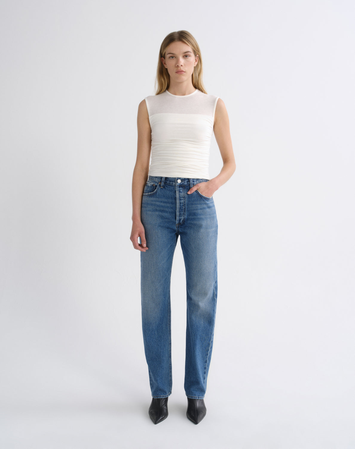 A model stands against a light gray studio background wearing a white sleeveless ribbed top and blue straight-leg jeans with black pointed shoes.