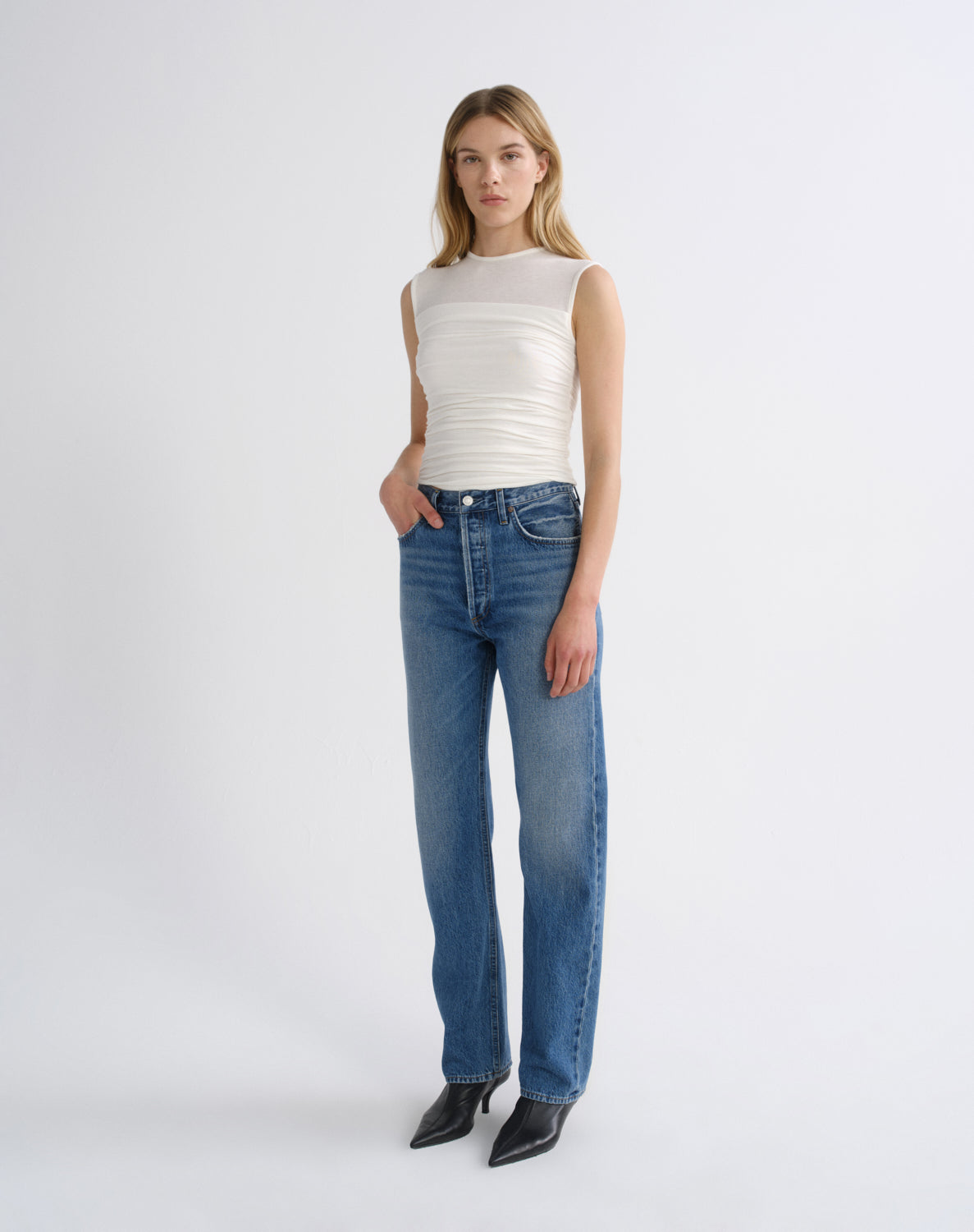 A model stands against a light gray studio background wearing a sleeveless cream ribbed top, blue straight-leg jeans, and black pointed shoes.