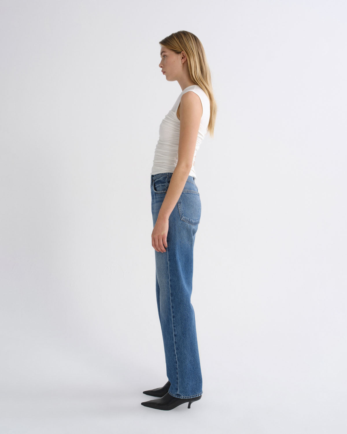 A woman in profile wearing a white ribbed tank top, blue straight-leg jeans, and black pointed-toe heels, standing against a plain light gray studio background.