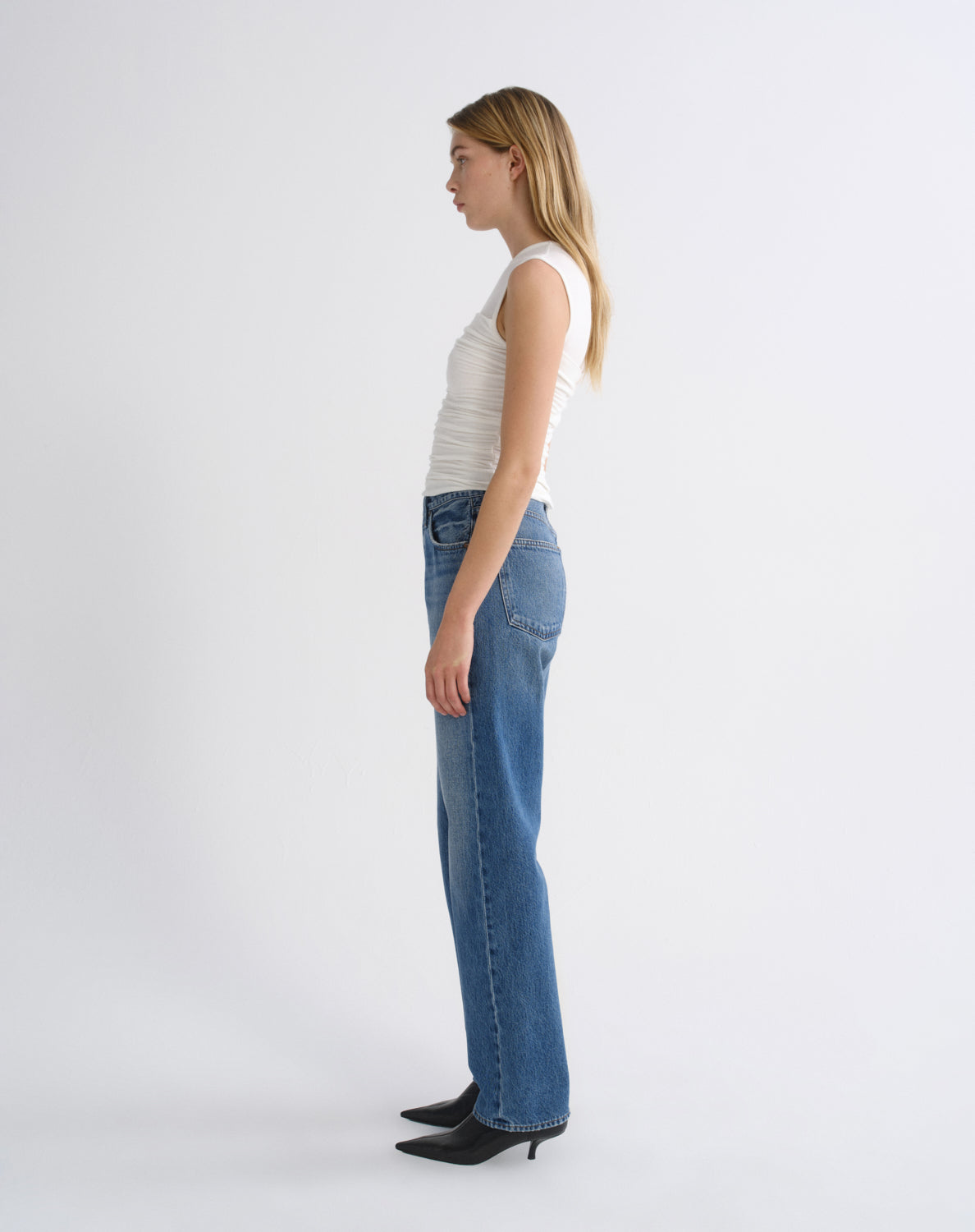 A woman in profile wearing a white ribbed tank top, blue straight-leg jeans, and black pointed-toe heels, standing against a plain light gray studio background.