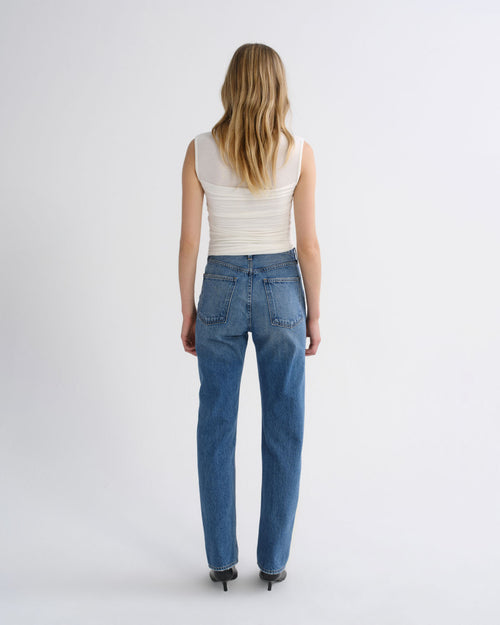 A woman viewed from behind wearing blue straight-leg jeans, a cream ribbed sleeveless top, and black pointed-toe heels against a plain light gray studio background.
