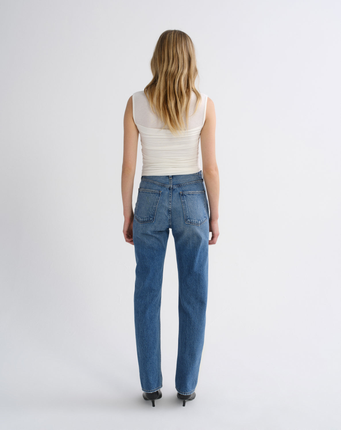 A woman viewed from behind wearing a cream ribbed sleeveless top and blue straight-leg jeans, standing against a plain light gray studio background.