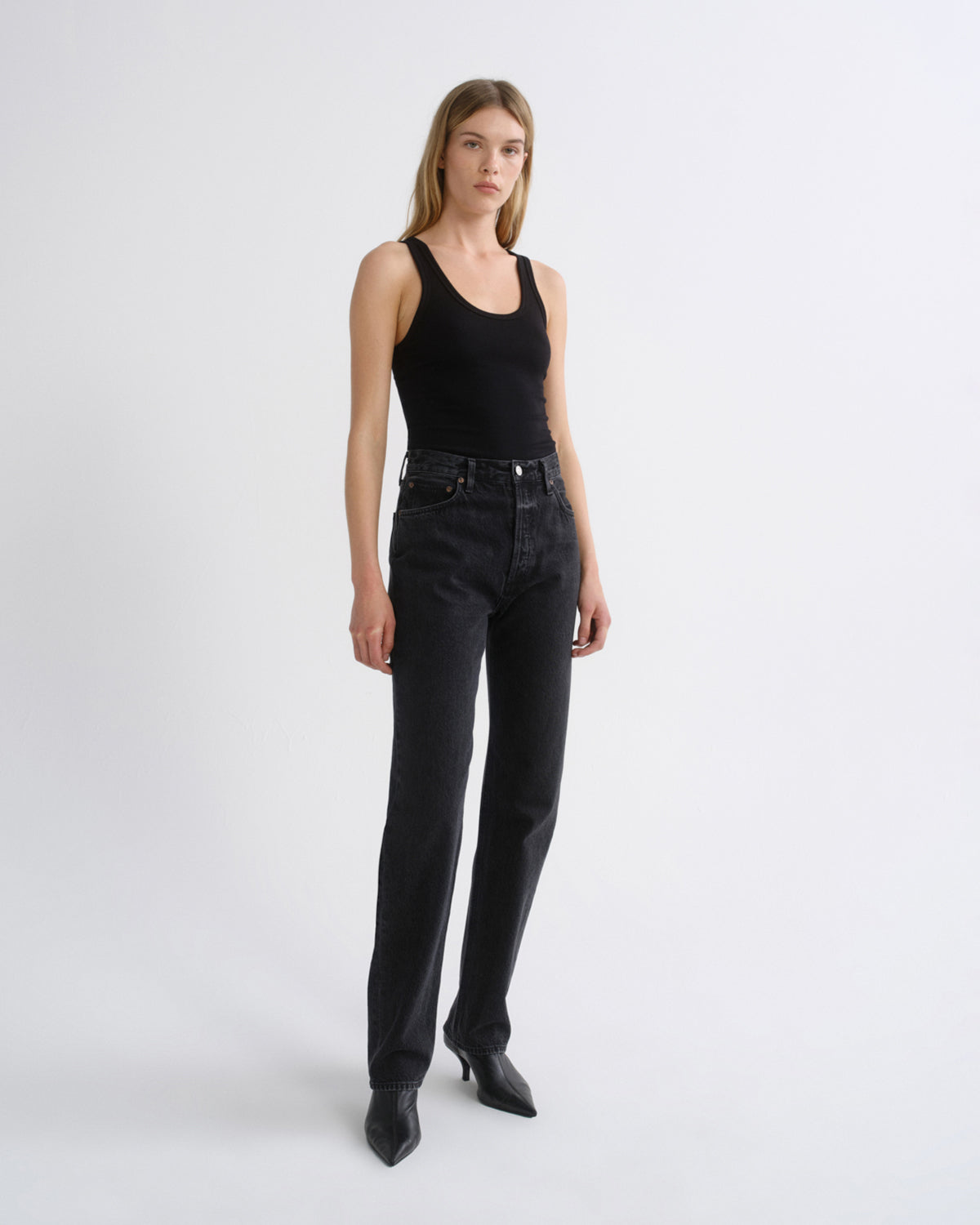 Woman wearing a black tank top with black straight leg pants on a grey background.