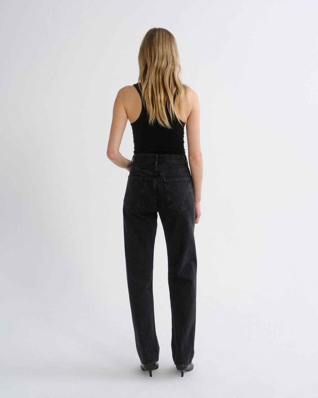 Woman wearing a black tank top with black straight leg pants viewed from the back on a grey background.