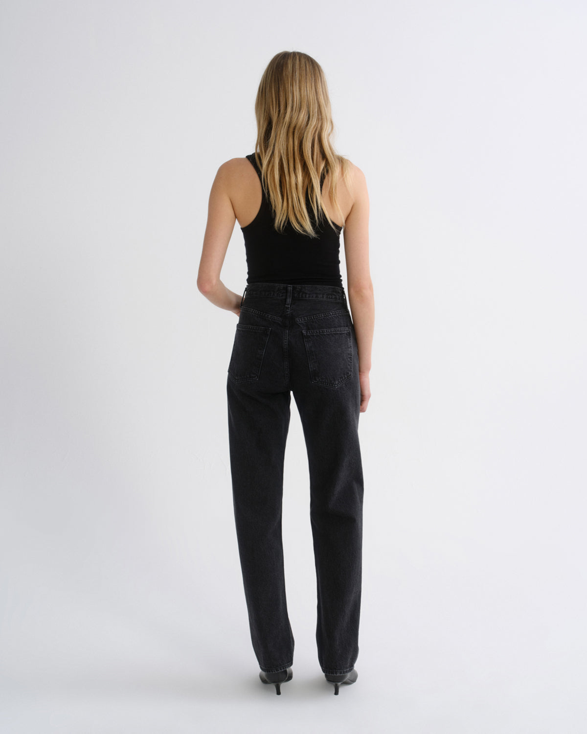 Woman wearing a black tank top with black straight leg pants viewed from the back on a grey background.