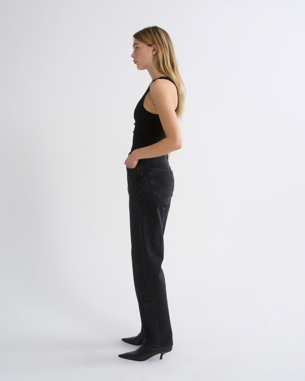 Woman wearing a black tank top with black straight leg pants viewed from the side on a grey background.