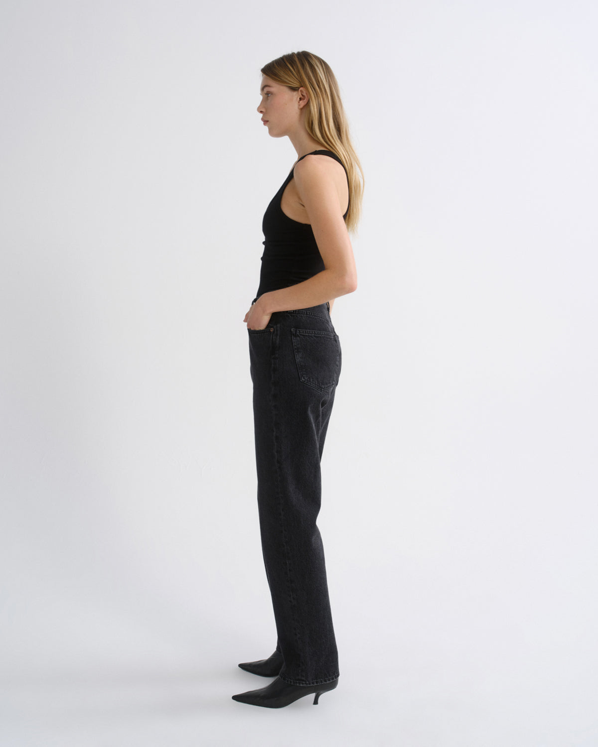 Woman wearing a black tank top with black straight leg pants viewed from the side on a grey background.