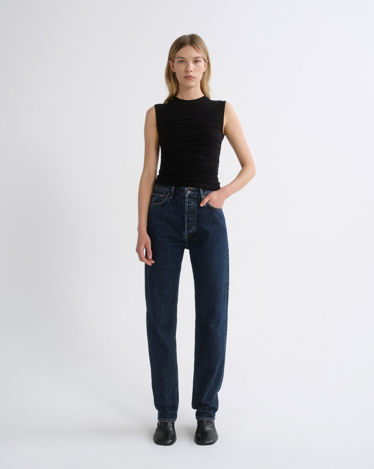 A model stands against a light gray studio background wearing a sleeveless black ribbed top, dark blue straight-leg jeans, and black shoes.