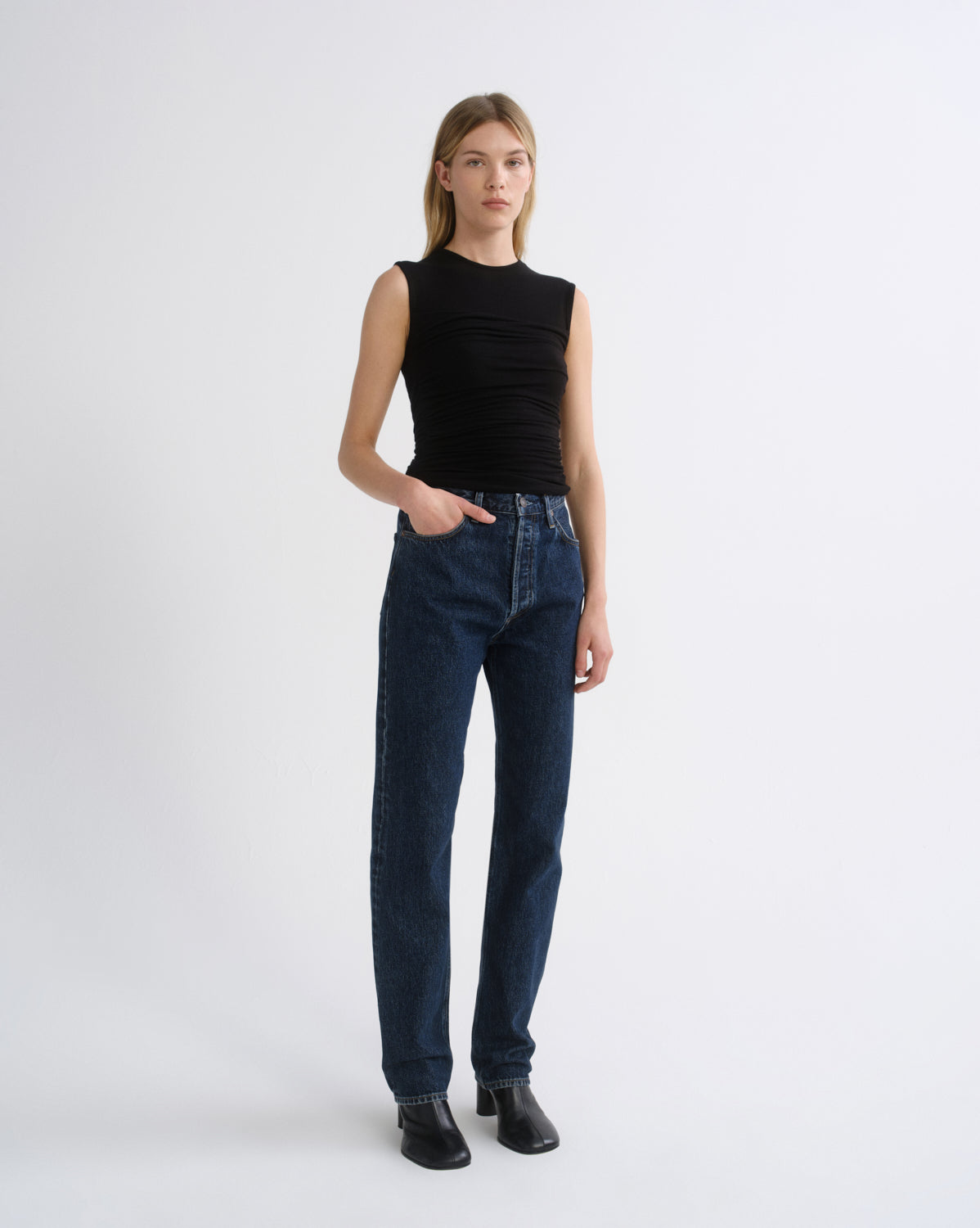 A model stands against a light gray studio background wearing a sleeveless black top, dark blue straight-leg jeans, and black ankle boots.