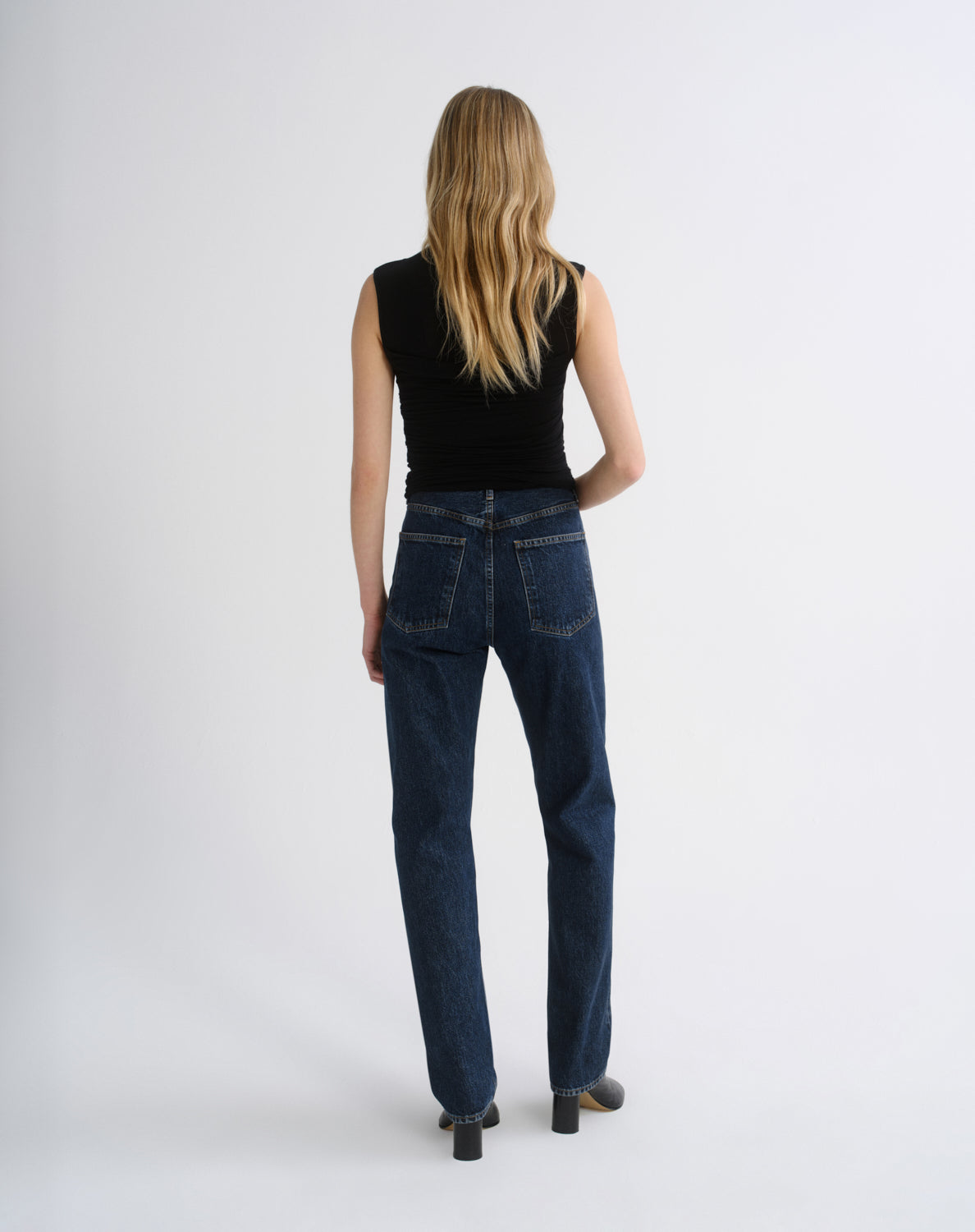 A woman viewed from behind wearing dark blue straight-leg jeans, a black sleeveless top, and black heels against a plain light gray studio background.