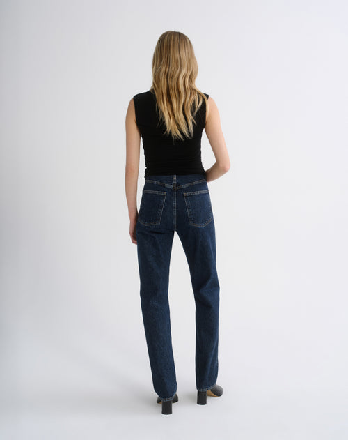 A woman viewed from behind wearing dark blue straight-leg jeans, a black sleeveless top, and black heels against a plain light gray studio background.