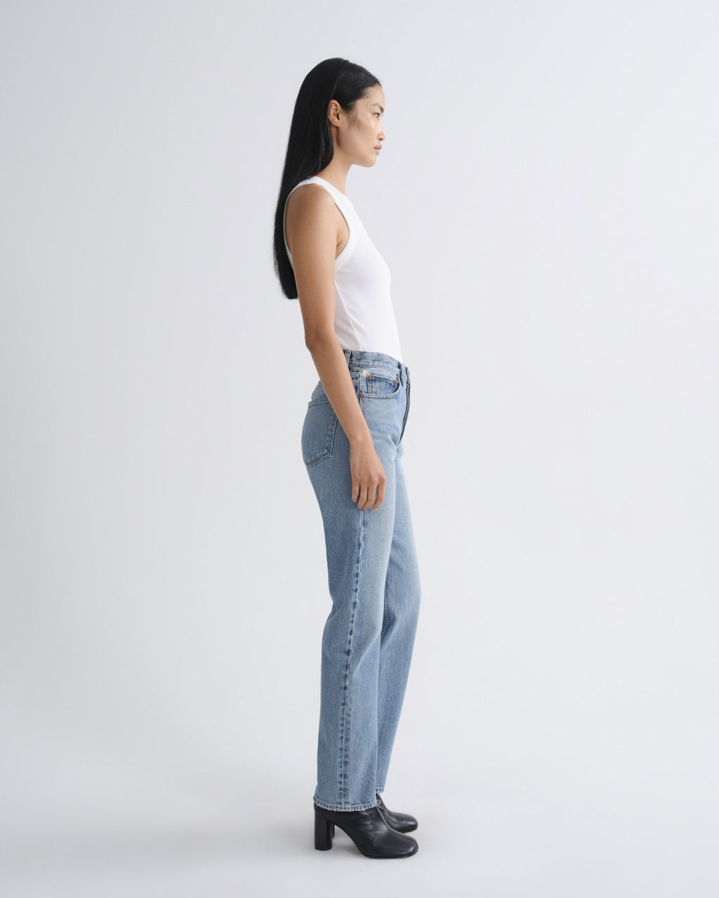 A woman in a white sleeveless top and light-wash blue jeans stands in profile against a plain light gray studio background, wearing black heeled boots.