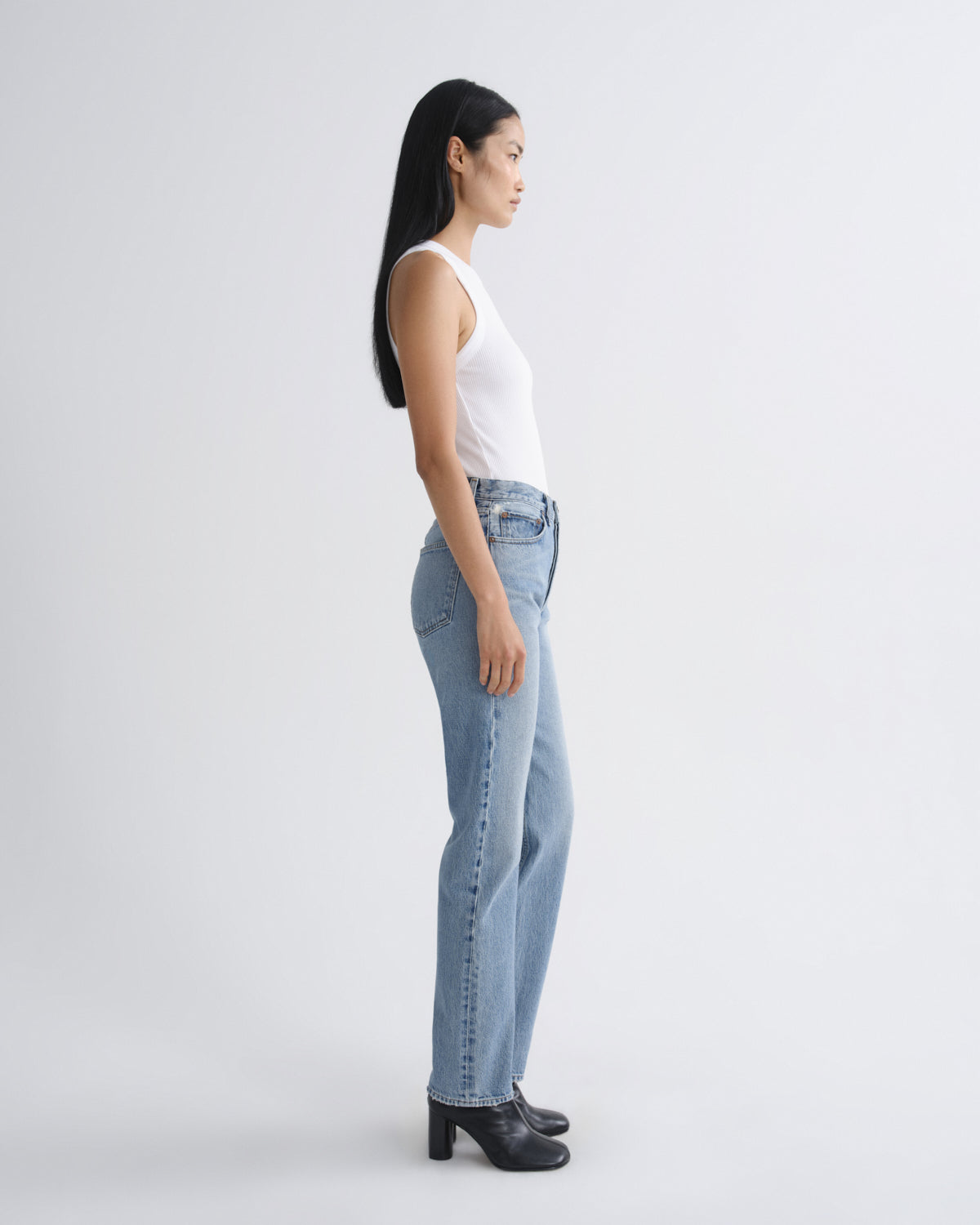 A woman in a white sleeveless top and light-wash blue jeans stands in profile against a plain light gray studio background, wearing black heeled boots.