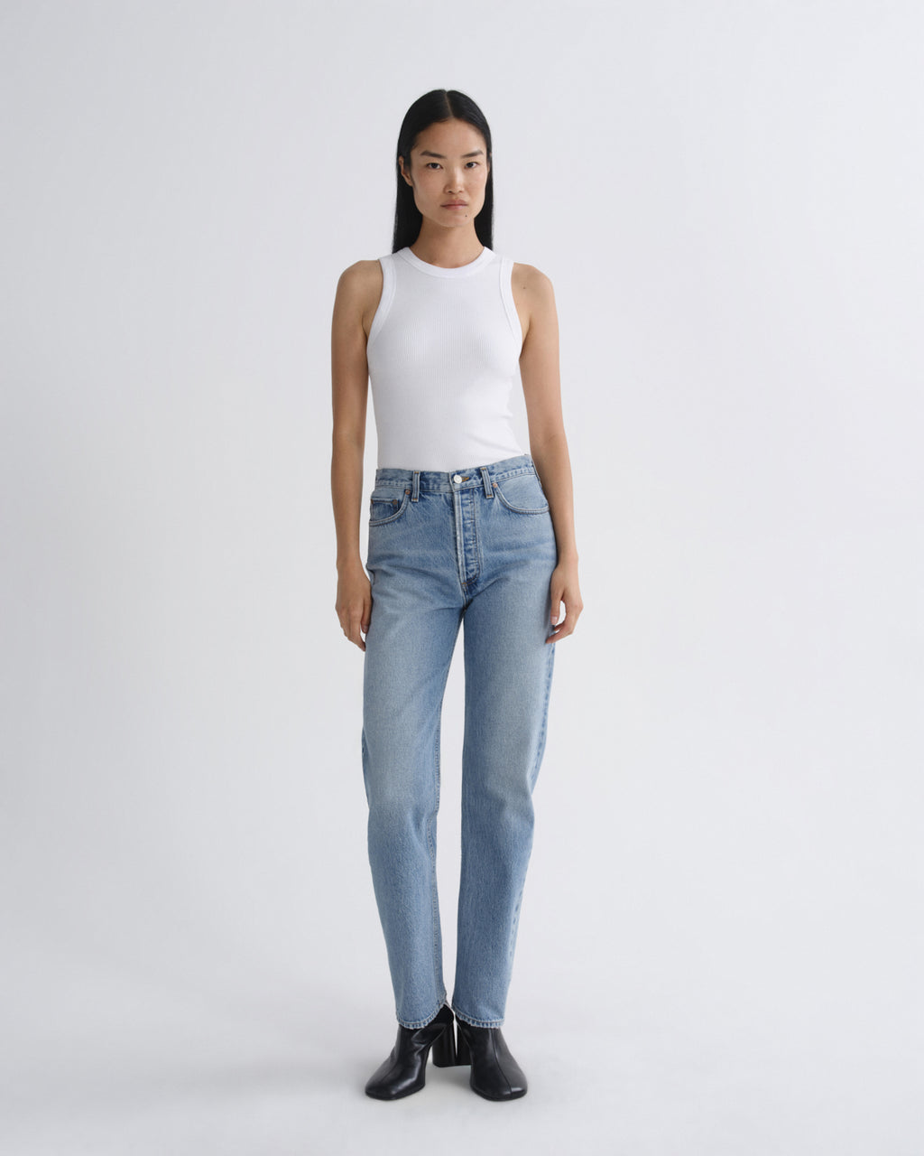 A model stands against a light gray studio background wearing a white sleeveless top, light-wash blue jeans, and black ankle boots.