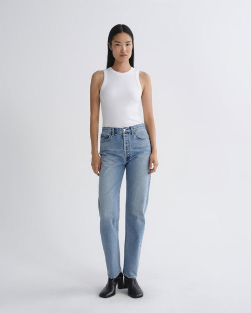 A model stands against a light gray studio background wearing a white sleeveless top, light-wash blue jeans, and black ankle boots.