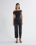 A model stands against a light gray studio background wearing a dark brown off-the-shoulder ruched top, dark gray wide-leg cropped jeans, and black strappy high-heeled sandals.