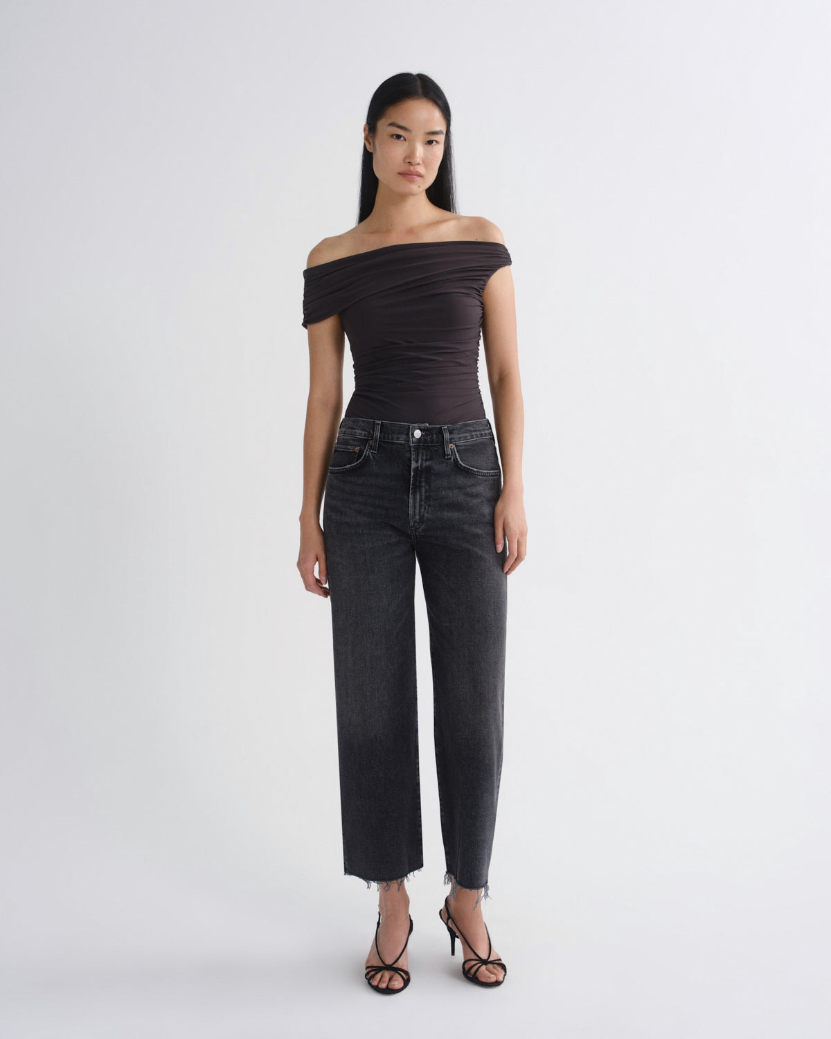 A model stands against a light gray studio background wearing a dark brown off-the-shoulder ruched top, dark gray wide-leg cropped jeans, and black strappy high-heeled sandals.