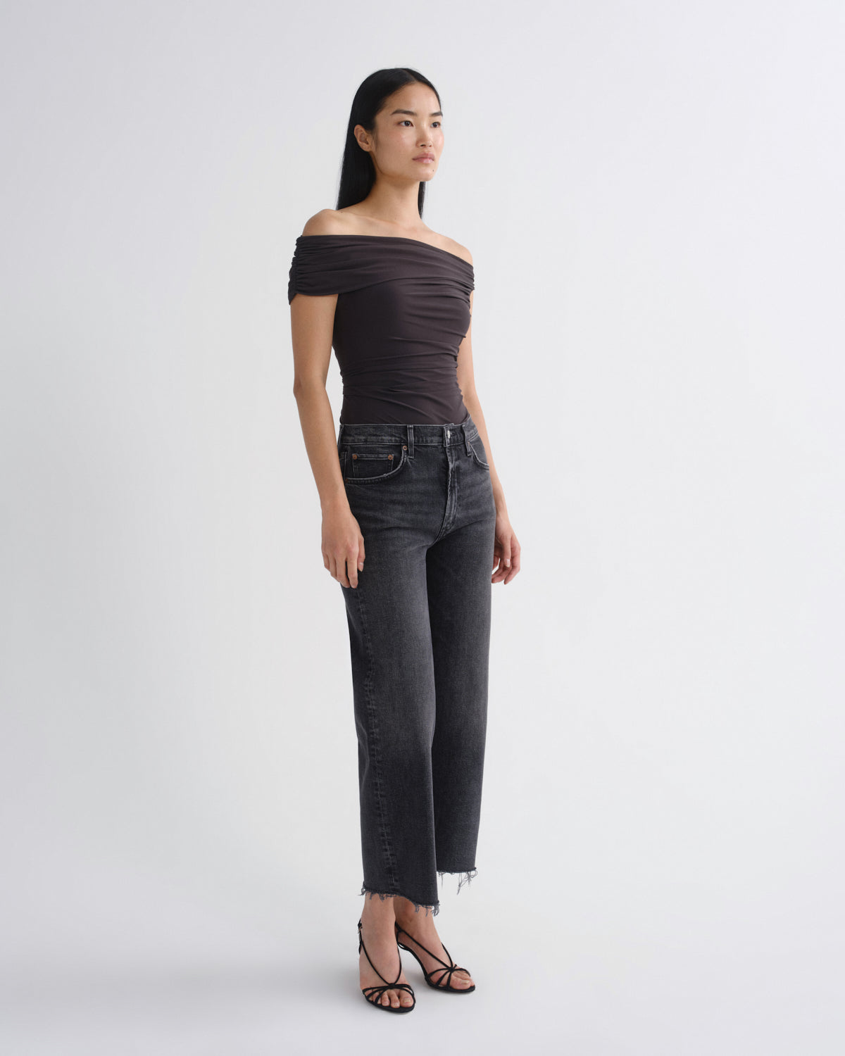 A woman modeling an off-the-shoulder dark brown ruched top and dark gray cropped jeans with frayed hems, paired with black strappy heels, against a plain light gray studio background.