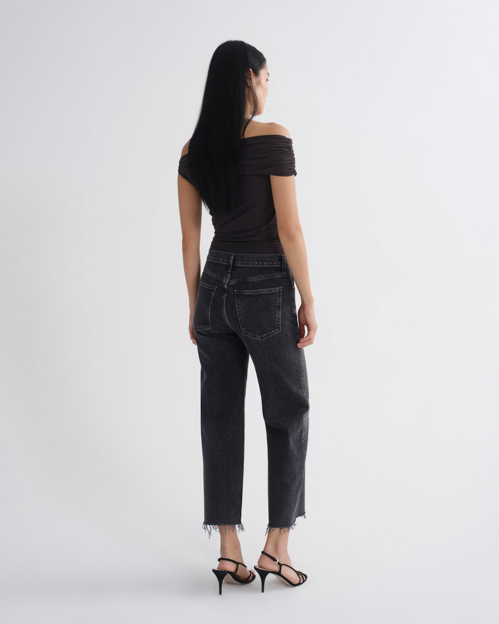 A woman shown from the back wearing a black off-the-shoulder top, dark gray cropped jeans with a raw hem, and black strappy high-heeled sandals, photographed against a light gray studio background.