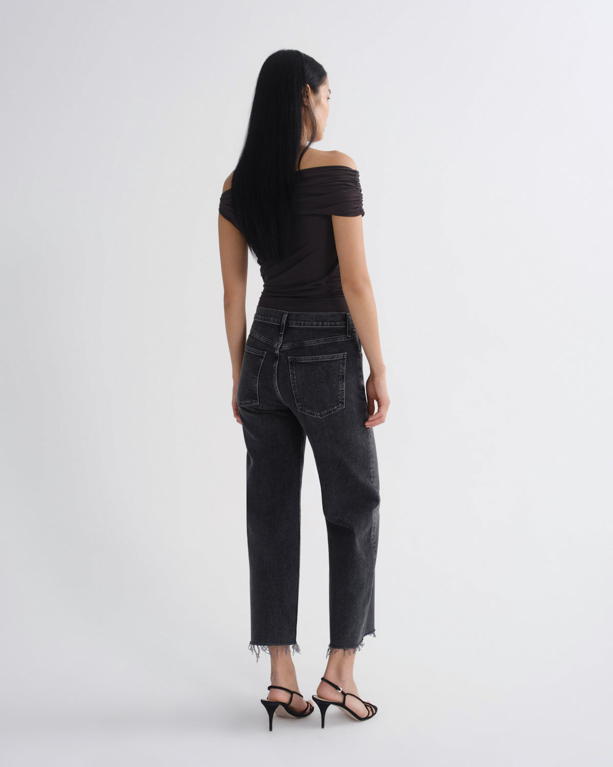 A woman shown from the back wearing a black off-the-shoulder top, dark gray cropped jeans with a raw hem, and black strappy high-heeled sandals, photographed against a light gray studio background.