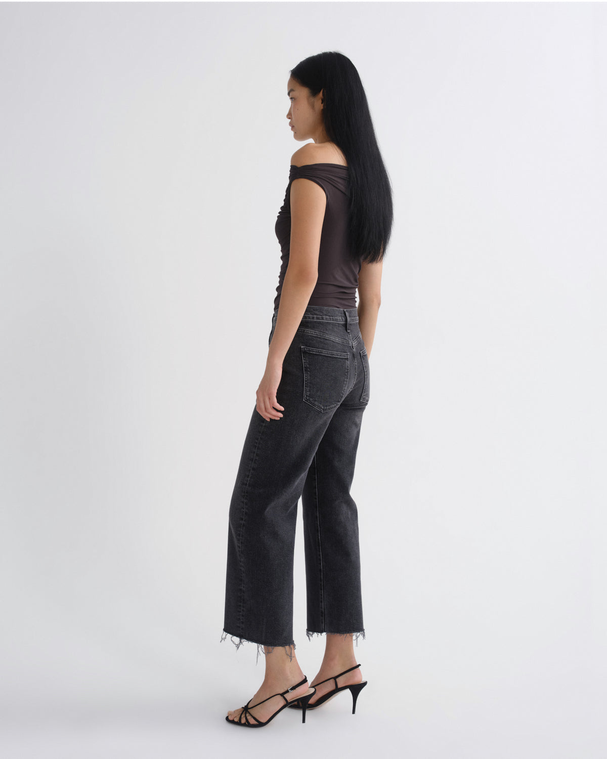A woman stands in profile wearing a dark brown off-the-shoulder top, dark gray cropped wide-leg jeans with raw hems, and black strappy high-heeled sandals against a plain light gray studio background.