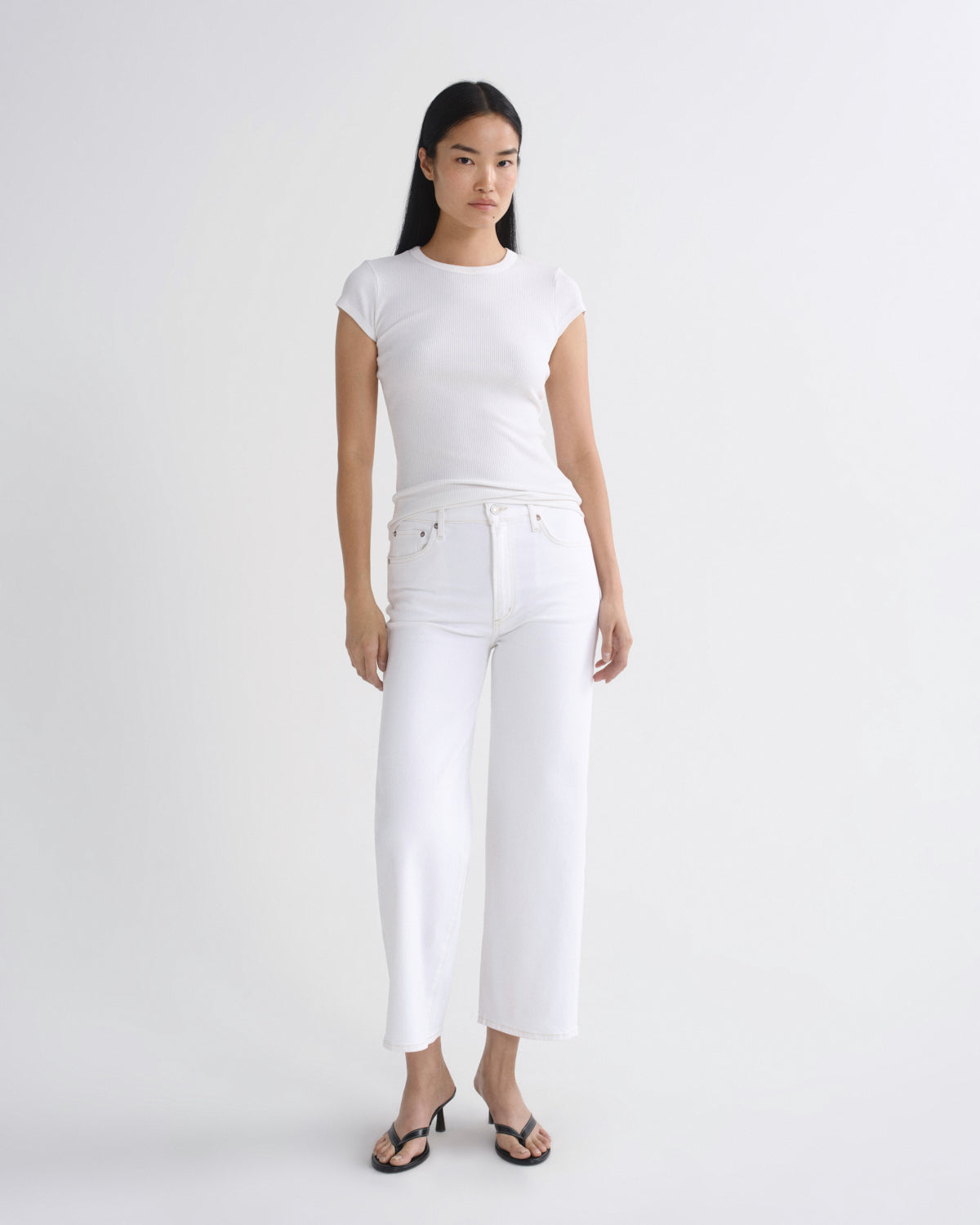A woman modeling a white short-sleeve crewneck t-shirt and white cropped jeans with black sandals, standing against a solid light gray studio background.