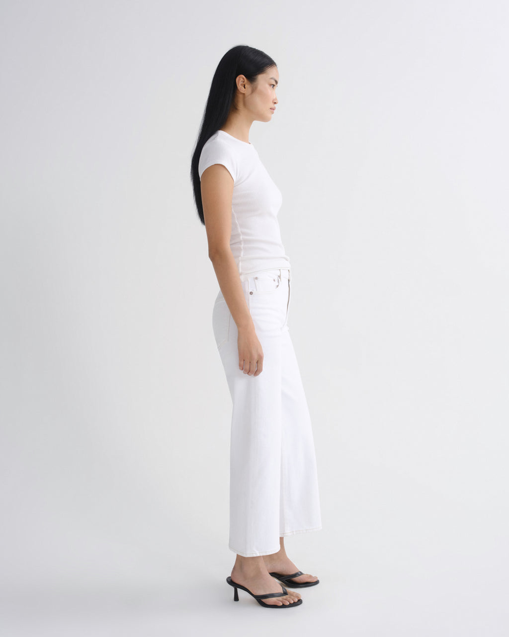 A woman in a side profile wearing a white short-sleeve top and white wide-leg cropped pants with black heeled sandals, photographed against a seamless light gray studio background.