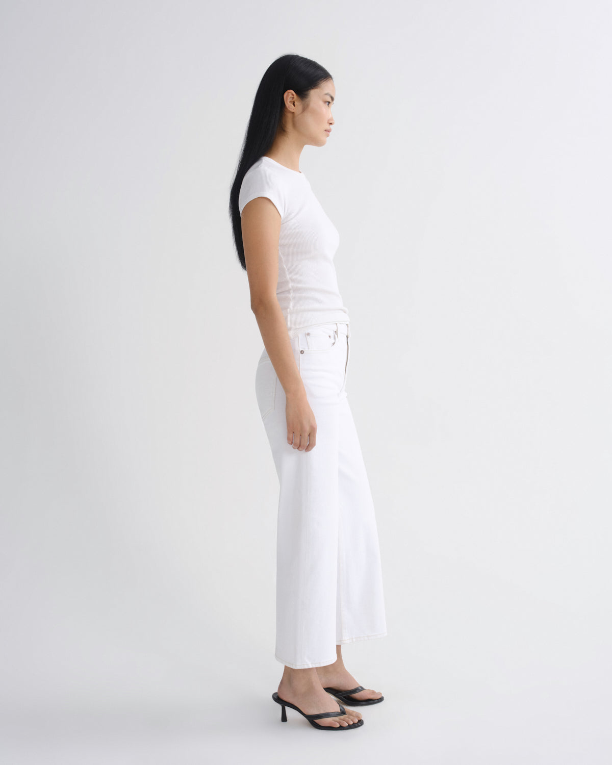 A woman in a side profile wearing a white short-sleeve top and white wide-leg cropped pants with black heeled sandals, photographed against a seamless light gray studio background.